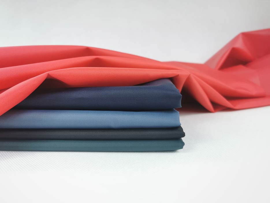 Zdjęcie przedstawiające czerwoną tkaninę wodoodporną ułożona swobodnie na materiałach w kolorze granatowym, niebieskim, czarnym oraz zielonym