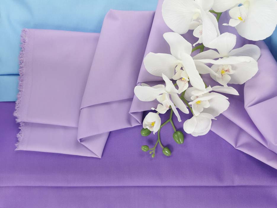 Zdjęcie przedstawiające jasnofioletową tkaninę Elanobawełnę Radus na tle materiałów w kolorze błękitnym oraz ciemnym fiolecie lawendowym