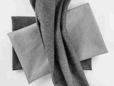 Zdjęcie przedstawiające tkaninę obiciową melanżową w odcieniu średniej szarości na tle materiałów w kontrastowych odcieniach