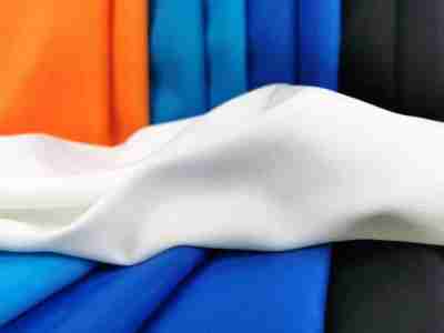 Zdjęcie prezentujące tkaninę diagonal w kolorze ciepłej bieli, ułożoną na tle materiałów w kolorze pomarańczowym, błękitnym, niebieskim i czarnym