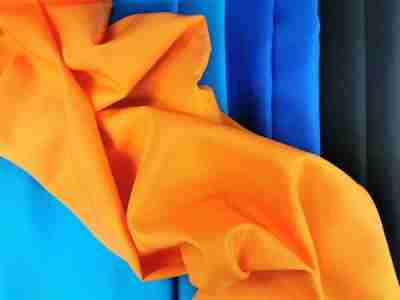 Zdjęcie przedstawiające pomarańczową tkaninę poliestrową Diagonal 1024 ułożoną swobodnie na materiałach w kolorze błękitnym, niebieskim oraz czarnym